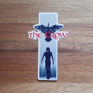 May include: A sticker featuring a black and white image of a crow with the text "The Crow" in red. The crow is flying with its wings spread wide. Below the crow is a black and white image of a person in a dark outfit walking towards the viewer.