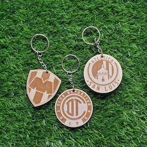 May include: Three wooden keychains with different designs. The first keychain features a brown and white shield with the letter "M" in the center. The second keychain features a brown circle with the text "CLUB DEPORTIVO AC" and the letters "DT" in the center. The third keychain features a brown circle with the text "ATLETICO SAN LUIS" and a building in the center.