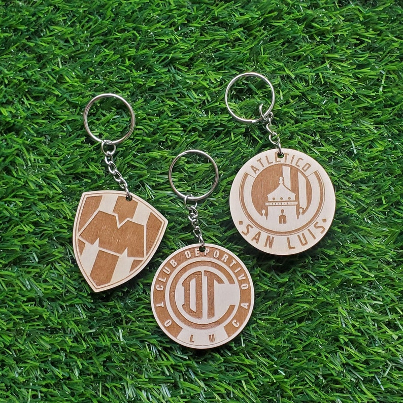 May include: Three wooden keychains with different designs. The first keychain features a brown and white shield with the letter "M" in the center. The second keychain features a brown circle with the text "CLUB DEPORTIVO AC" and the letters "DT" in the center. The third keychain features a brown circle with the text "ATLETICO SAN LUIS" and a building in the center.