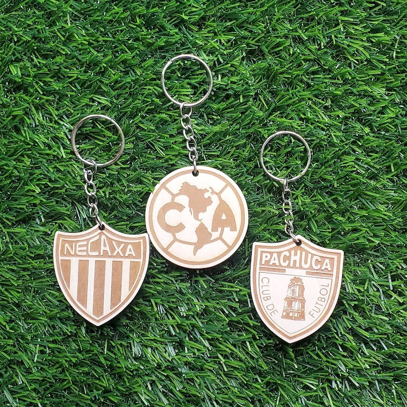 May include: Three wooden keychains with different soccer team logos. The keychains are brown and feature the logos for Club de Futbol Pachuca, Club America, and Club Necaxa.