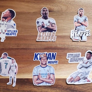 May include: Six different stickers featuring a soccer player with the name "Kylian Mbappe" printed on each sticker. The stickers are printed on white paper with blue and gold lettering.