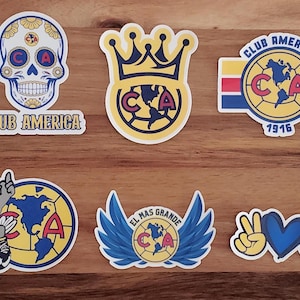 May include: A collection of Club America stickers featuring various designs. Includes a sugar skull, a crown, the team logo with the year 1916, a soccer eagle, a logo with wings, and a peace sign with a heart.