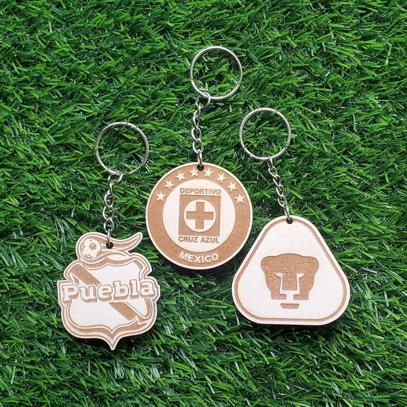 May include: Three wooden keychains with different designs. The first keychain features a brown and white shield with the word "Puebla" on it. The second keychain is round and features a white cross on a blue background with the text "Deportivo Cruz Azul Mexico". The third keychain is a triangle with a brown and white puma head on it.