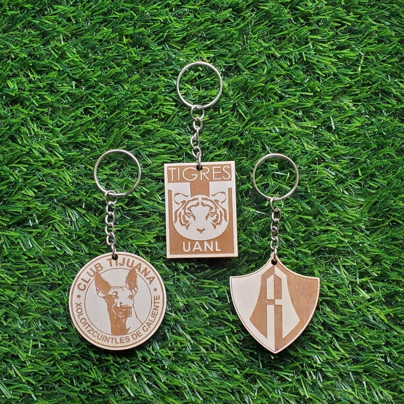 May include: Three wooden keychains with different designs. The first keychain features a brown and white dog with the text "CLUB TIJUANA XOLOITZCUINTLES DE CALIENTE". The second keychain features a tiger with the text "TIGRES UANL". The third keychain features a shield with the letter "A" inside.