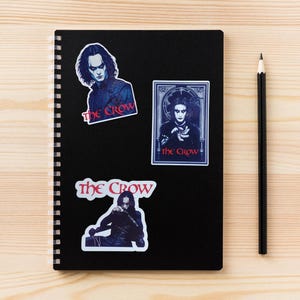 May include: A black notebook with a spiral binding has three stickers on the front. The stickers feature the character Eric Draven from the movie "The Crow". The stickers have the text "The Crow" in red and white.