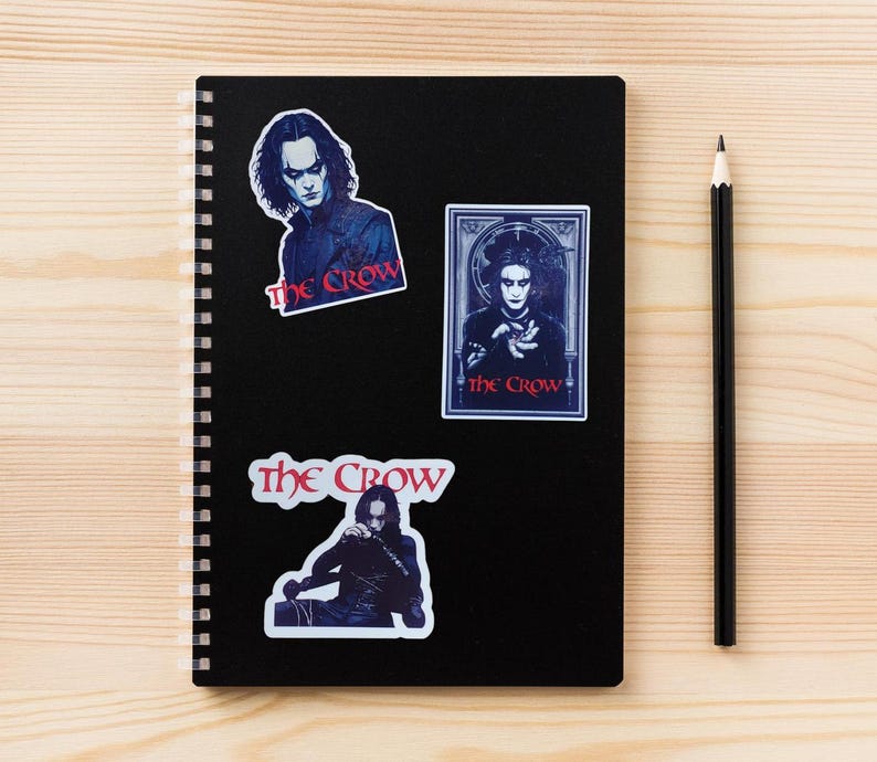 May include: A black notebook with a spiral binding has three stickers on the front. The stickers feature the character Eric Draven from the movie "The Crow". The stickers have the text "The Crow" in red and white.