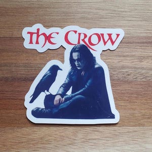 May include: A white sticker with the words "The Crow" in red and blue lettering. The sticker features a black and white image of a man with black hair and a black crow sitting beside him.