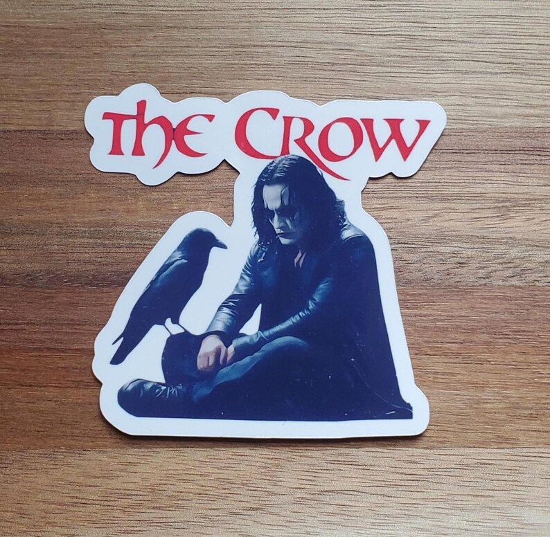 May include: A white sticker with the words "The Crow" in red and blue lettering. The sticker features a black and white image of a man with black hair and a black crow sitting beside him.