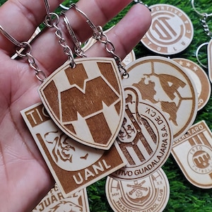 May include: A collection of wooden keychains featuring different sports team logos. The keychains are made of light brown wood and have a variety of designs, including a tiger, a shield with a letter M, and a soccer ball. The keychains are attached to silver key rings.