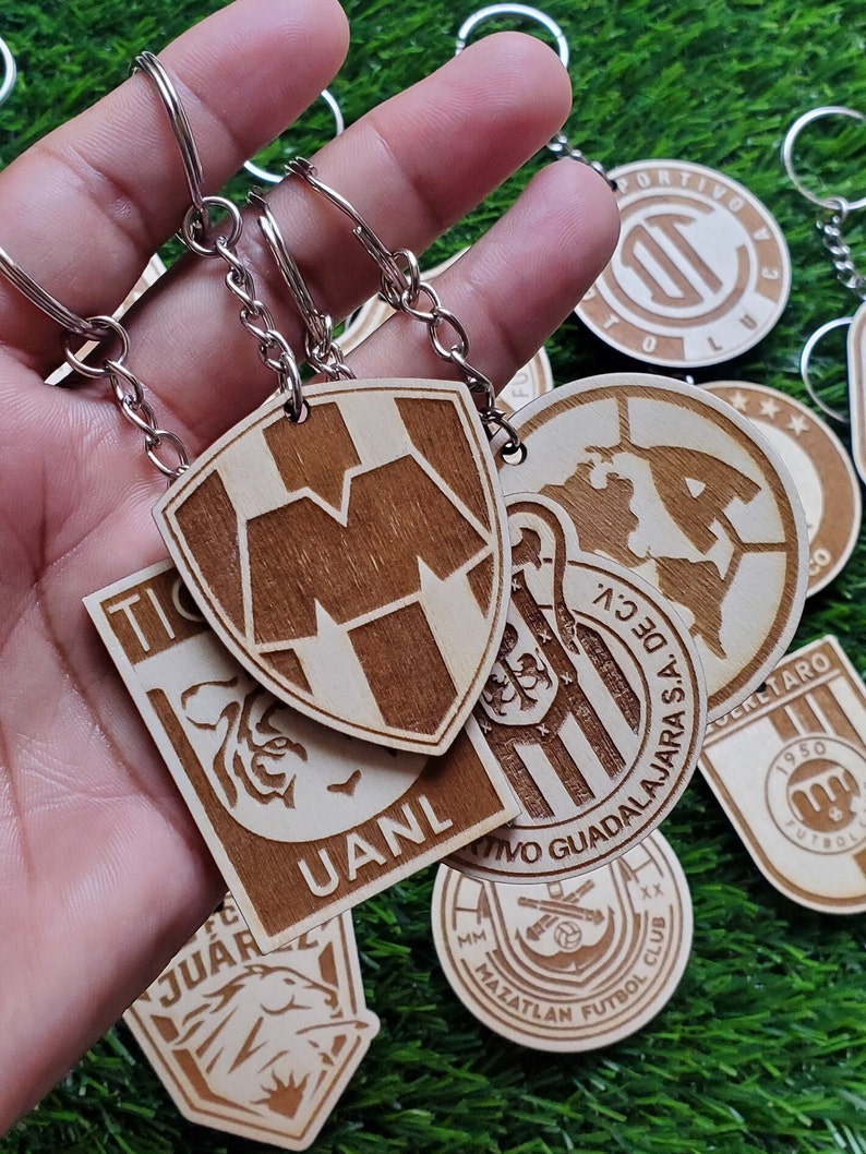 May include: A collection of wooden keychains featuring different sports team logos. The keychains are made of light brown wood and have a variety of designs, including a tiger, a shield with a letter M, and a soccer ball. The keychains are attached to silver key rings.