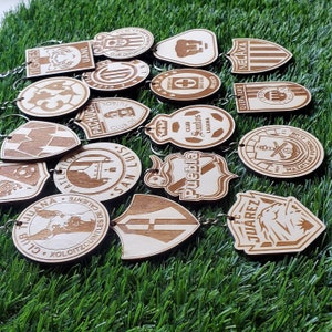 May include: A collection of wooden keychains featuring logos of different Mexican soccer teams. The keychains are made of light brown wood and have a variety of designs, including team names, crests, and mascots.