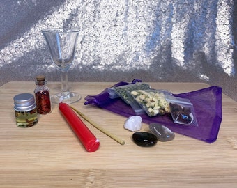 May include: A purple pouch with a variety of herbs, crystals, and other items for a spell or ritual. The pouch is on a wooden surface with a glass goblet, a red candle, a small bottle of oil, and a small bottle of glitter.