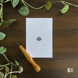 Può includere: Una carta bianca con un timbro di zucca grigia, uno strumento con manico in legno e viti a foglie verdi su una superficie di legno scuro. La carta è centrata, con lo strumento e le viti che incorniciano la scena. Il timbro è una zucca stilizzata.