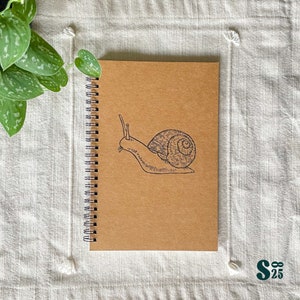 Pode incluir: Um caderno espiral marrom com uma ilustração em preto e branco de um caracol na capa.