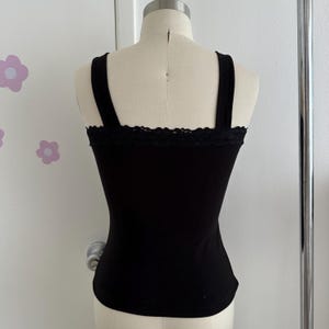 May include: Black tank top with wide straps and a lace trim along the neckline. The fitted top is displayed on a mannequin. The background is a white wall with flower decals.