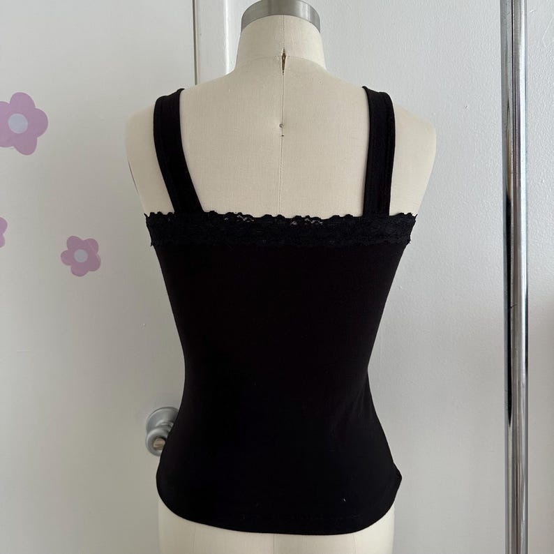 May include: Black tank top with wide straps and a lace trim along the neckline. The fitted top is displayed on a mannequin. The background is a white wall with flower decals.