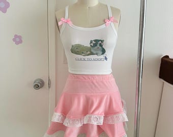 May include: A white ribbed tank top featuring a graphic of two kittens with pink bows and the text CLICK TO ADOPT, accented with small pink shoulder bows. Paired with a pink tiered mini skirt featuring white lace trim on each ruffled layer.
