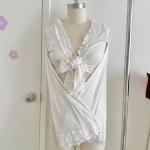 May include: A white long-sleeved top with a tie-front closure and lace trim. The top is displayed on a mannequin, showcasing its design. The neckline and hem feature delicate lace detailing, adding a touch of elegance to the garment.