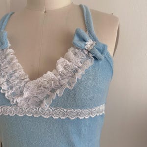May include: Light blue tank top with white lace trim and bows. The top features a ruffled lace neckline and a delicate lace trim at the waist. The bows are placed on the straps, adding a touch of elegance to the design.