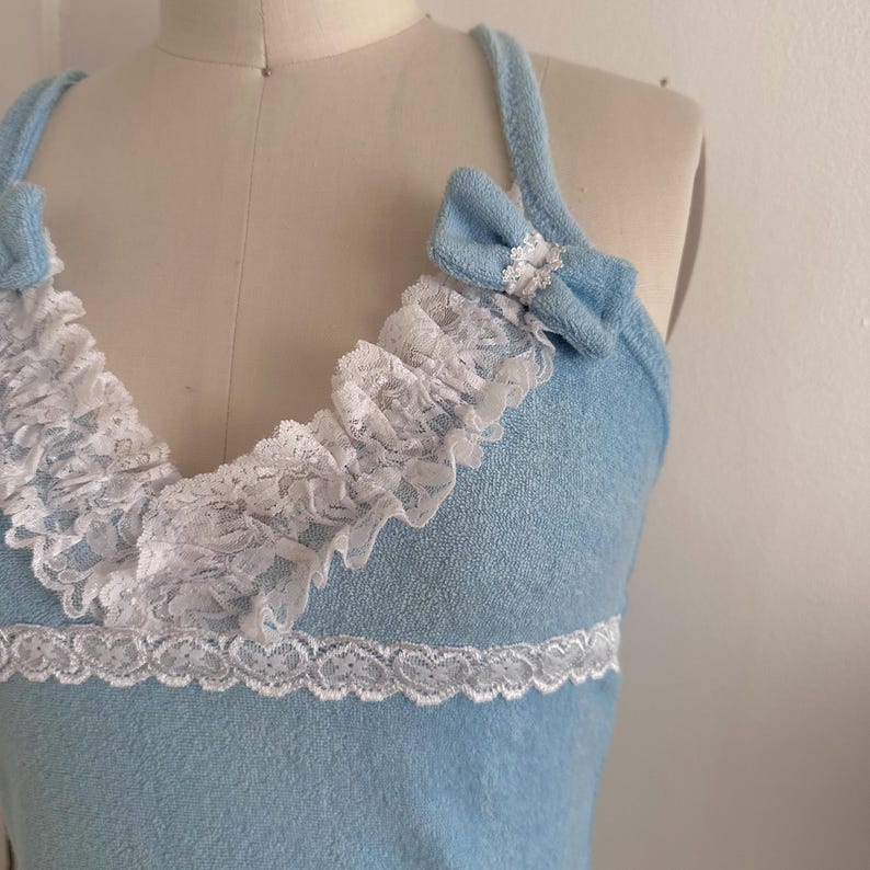 May include: Light blue tank top with white lace trim and bows. The top features a ruffled lace neckline and a delicate lace trim at the waist. The bows are placed on the straps, adding a touch of elegance to the design.