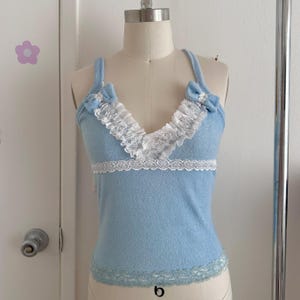 May include: A light blue camisole top with white lace trim and ruffles. The top features a V-neckline and small bows on the straps. The bottom hem and the area below the bust are also trimmed with white lace. The camisole is displayed on a mannequin.