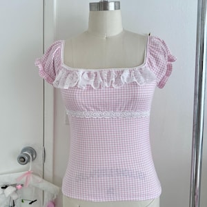 May include: A pink and white gingham top with a square neckline and short sleeves. The top has white lace trim on the neckline and waist. The garment is displayed on a mannequin.