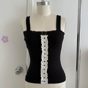 May include: A black tank top with wide straps and a square neckline. The top features a white lace trim along the center and the top edge. The tank top is displayed on a mannequin.