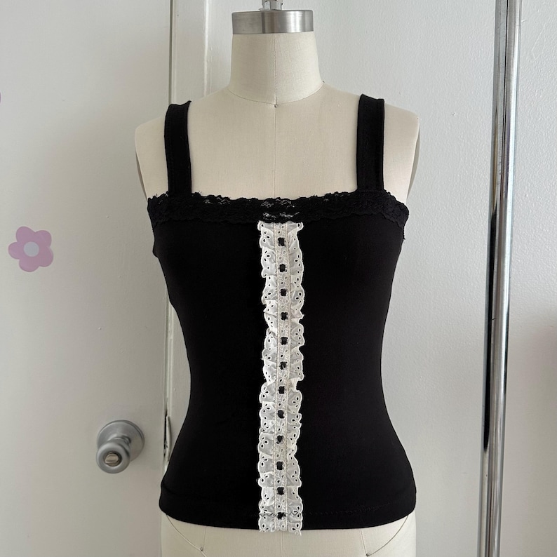 May include: A black tank top with wide straps and a square neckline. The top features a white lace trim along the center and the top edge. The tank top is displayed on a mannequin.