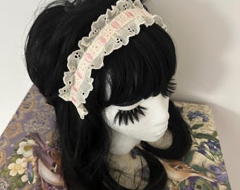 Può includere: Una fascia per capelli in pizzo bianco con dettagli rosa e un bordo smerlato. La fascia è indossata su una testa di manichino con capelli neri e lunghe ciglia. Lo sfondo presenta un motivo floreale.