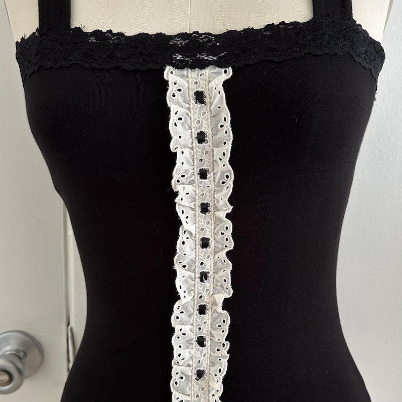 May include: A black tank top with a white eyelet trim down the center and black lace trim along the top edge. The tank top has thin straps and a fitted silhouette.