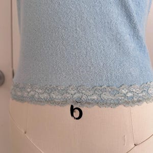 May include: Light blue sleeveless top with a textured terry cloth fabric. The bottom edge features a delicate, light blue lace trim. The top is displayed on a mannequin, showcasing its shape and design.