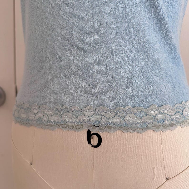 May include: Light blue sleeveless top with a textured terry cloth fabric. The bottom edge features a delicate, light blue lace trim. The top is displayed on a mannequin, showcasing its shape and design.