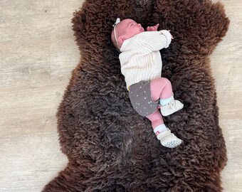 May include: A newborn baby wearing a cream-colored sweater, gray shorts with star patterns, and white booties lies on a dark brown sheepskin rug. The baby has a white headband with a bow.