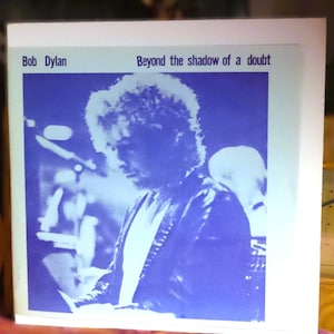 Può includere: Una copertina quadrata per disco in vinile con una fotografia monocromatica blu e bianca di Bob Dylan durante un'esibizione. Il testo Bob Dylan e Beyond the shadow of a doubt è stampato in nero sul bordo bianco che circonda l'immagine.