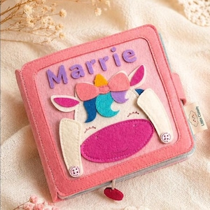 May include: A pink felt book with the name "Marrie" on the cover. The cover features a unicorn design with a pink mouth, white ears, and a colourful bow. The book is closed with a small red button.
