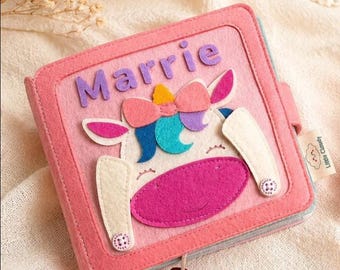 May include: A pink felt book with the name "Marrie" on the cover. The cover features a unicorn design with a pink mouth, white ears, and a colorful bow. The book is closed with a small red button.