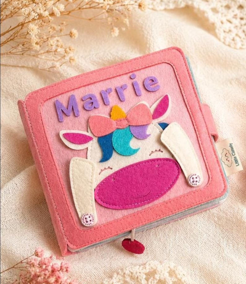 May include: A pink felt book with the name "Marrie" on the cover. The cover features a unicorn design with a pink mouth, white ears, and a colourful bow. The book is closed with a small red button.