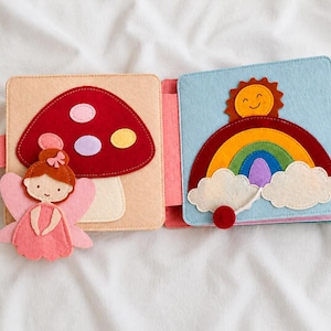 May include: Open felt book featuring a fairy, mushroom, rainbow, and sun design. The fairy is pink with orange hair. The mushroom is red with white, purple, and yellow spots. The rainbow has multiple colours.