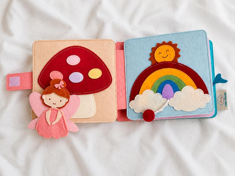 May include: Open felt book featuring a fairy, mushroom, rainbow, and sun design. The fairy is pink with orange hair. The mushroom is red with white, purple, and yellow spots. The rainbow has multiple colours.