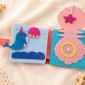 May include: A colourful felt book with an ocean theme. The open pages display a blue narwhal, a pink jellyfish, and a pink seashell with a starfish. The book is designed with bright colours and tactile elements for children.