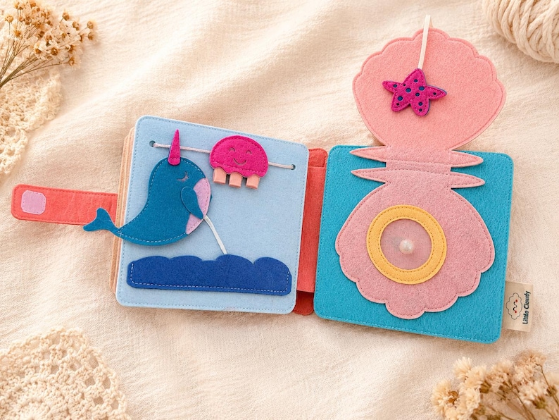 May include: A colourful felt book with an ocean theme. The open pages display a blue narwhal, a pink jellyfish, and a pink seashell with a starfish. The book is designed with bright colours and tactile elements for children.