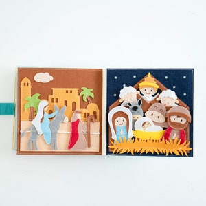 May include: A felt book with two open pages depicting a Nativity scene. One page shows Mary and Joseph on a donkey, the other shows the baby Jesus in a manger with the three wise men and animals. The book has a teal clasp.