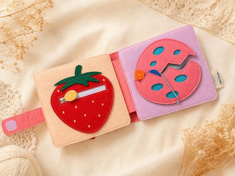 May include: A felt activity book, open to reveal a strawberry and a cracked egg. The strawberry has a zip, and the egg features buttons and string. The book is made of red, pink, green, and beige felt.