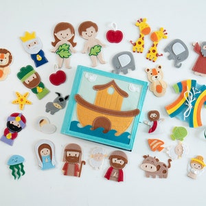 May include: A felt book with an ark design, surrounded by felt figures of animals, people, and religious symbols. The figures include giraffes, lions, and people from a biblical story. The book and figures are in various colors.