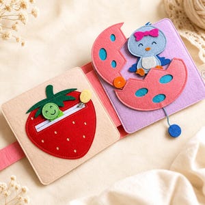 May include: A colourful felt activity book for babies. The open book displays a red strawberry with a zip and a smiling green caterpillar. The right page shows a blue bird emerging from a pink egg. The book is on a cream-coloured surface.