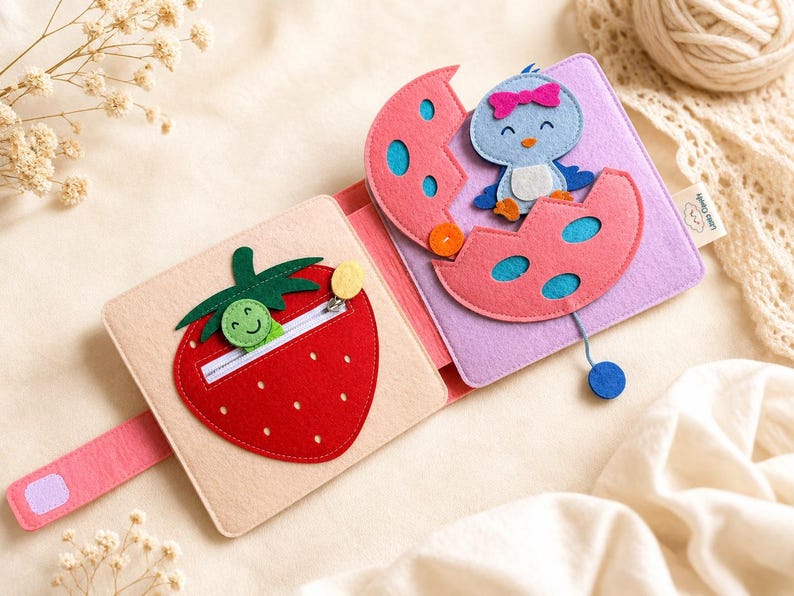May include: A colourful felt activity book for babies. The open book displays a red strawberry with a zip and a smiling green caterpillar. The right page shows a blue bird emerging from a pink egg. The book is on a cream-coloured surface.