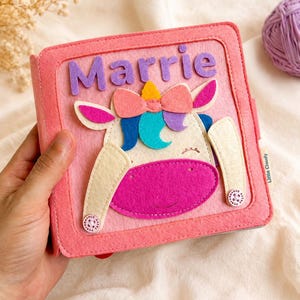 May include: A pink felt book featuring a unicorn design and the name "Marrie" in purple letters. The unicorn has a pink bow, blue and purple accents, and a yellow horn. The book is held in a hand.
