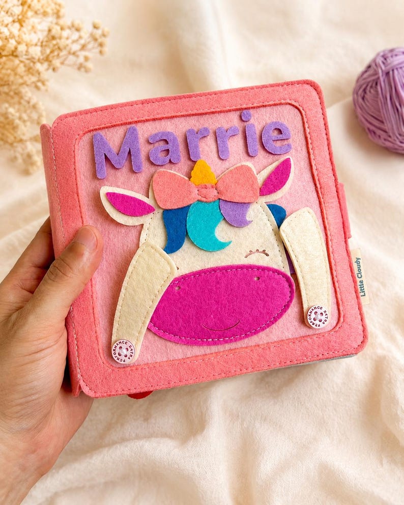 May include: A pink felt book featuring a unicorn design and the name "Marrie" in purple letters. The unicorn has a pink bow, blue and purple accents, and a yellow horn. The book is held in a hand.