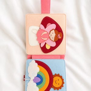 May include: A colourful felt activity book with a pink handle. The top section shows a pink fairy on a red heart. The bottom section has a rainbow, sun, and clouds. Designed for young children.