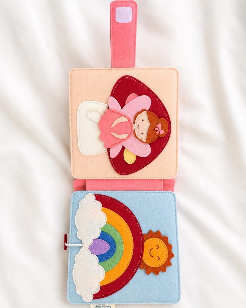 May include: A colourful felt activity book with a pink handle. The top section shows a pink fairy on a red heart. The bottom section has a rainbow, sun, and clouds. Designed for young children.
